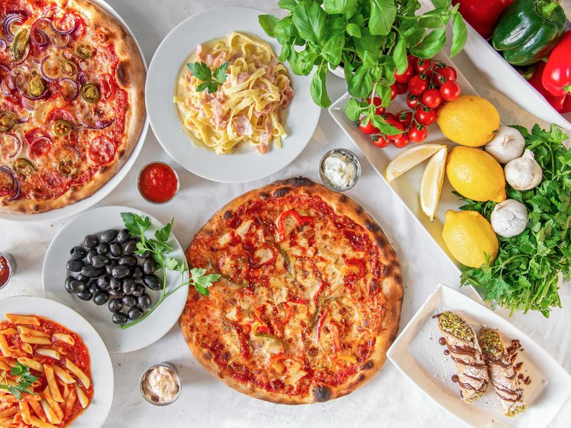 Overhead view of pizzas, pasta, olives and fresh vegetables and herbs